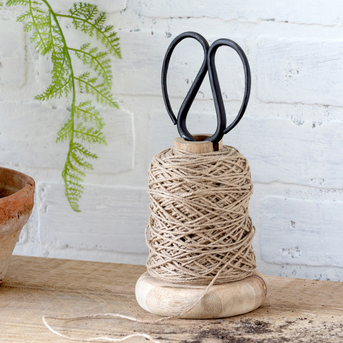 wood garden jute spool with black snip scissors tucked inside on a white brick wall with an artificial fern 