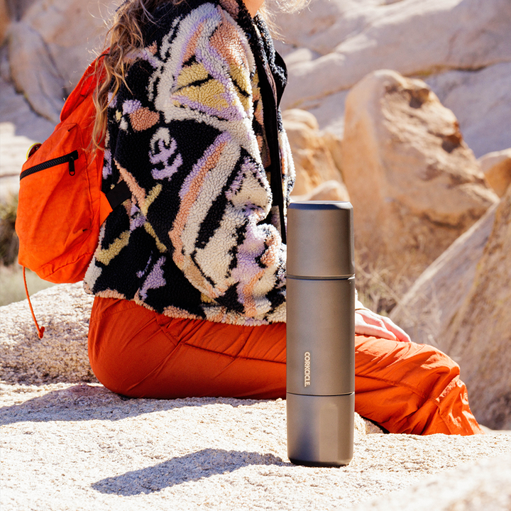 a female in a colorful jacket sitting on rock with grey Corkcicle thermos