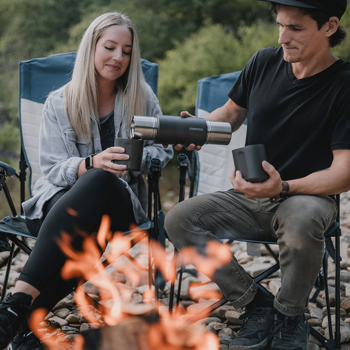 male and female hikers by fire pouring a cup of coffee from a Corkcicle brand thermos 
