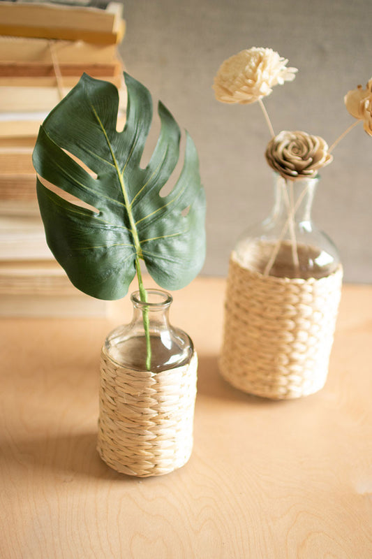 two seagrass wrapped clear vases with monstera leaf and wood flowers 